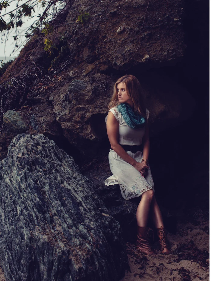 A woman in a white dress and a teal scarf standing among dark rocks