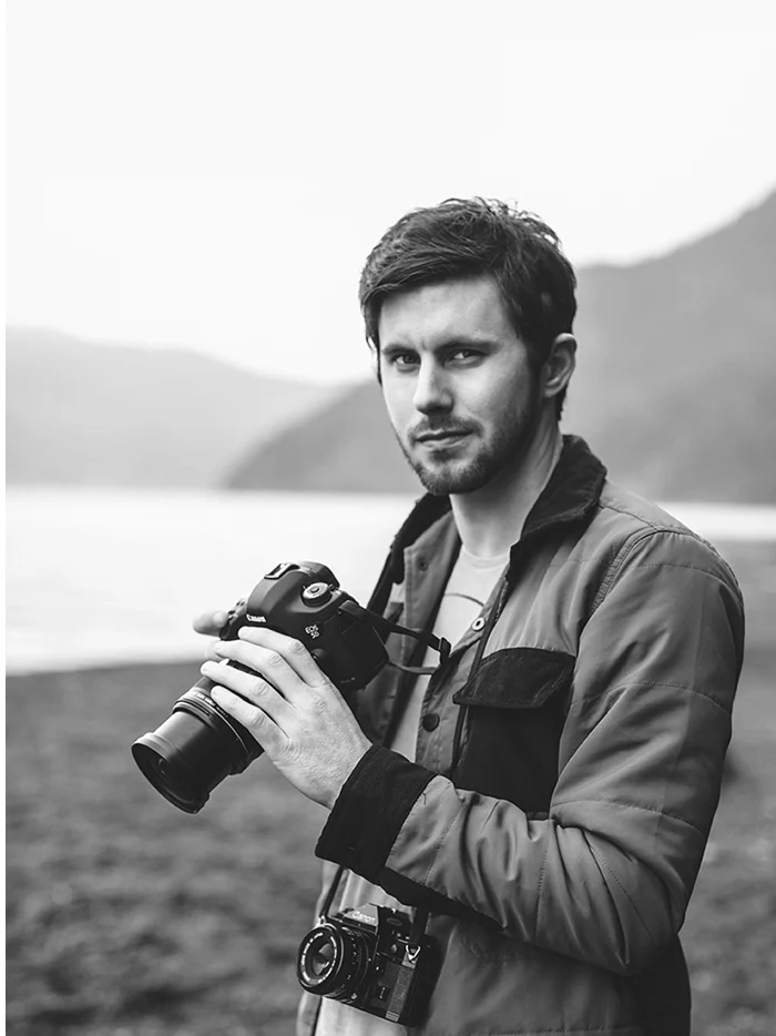 A man in a gray jacket holding a camera on the shore of a misty lake