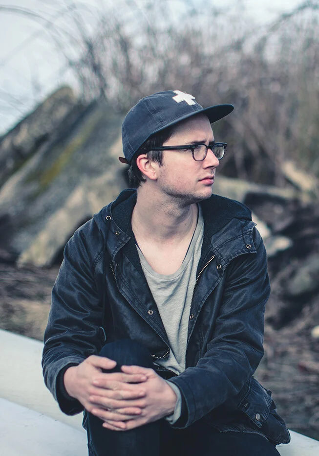 A young man in a cap and glasses sitting on rocks by the lake