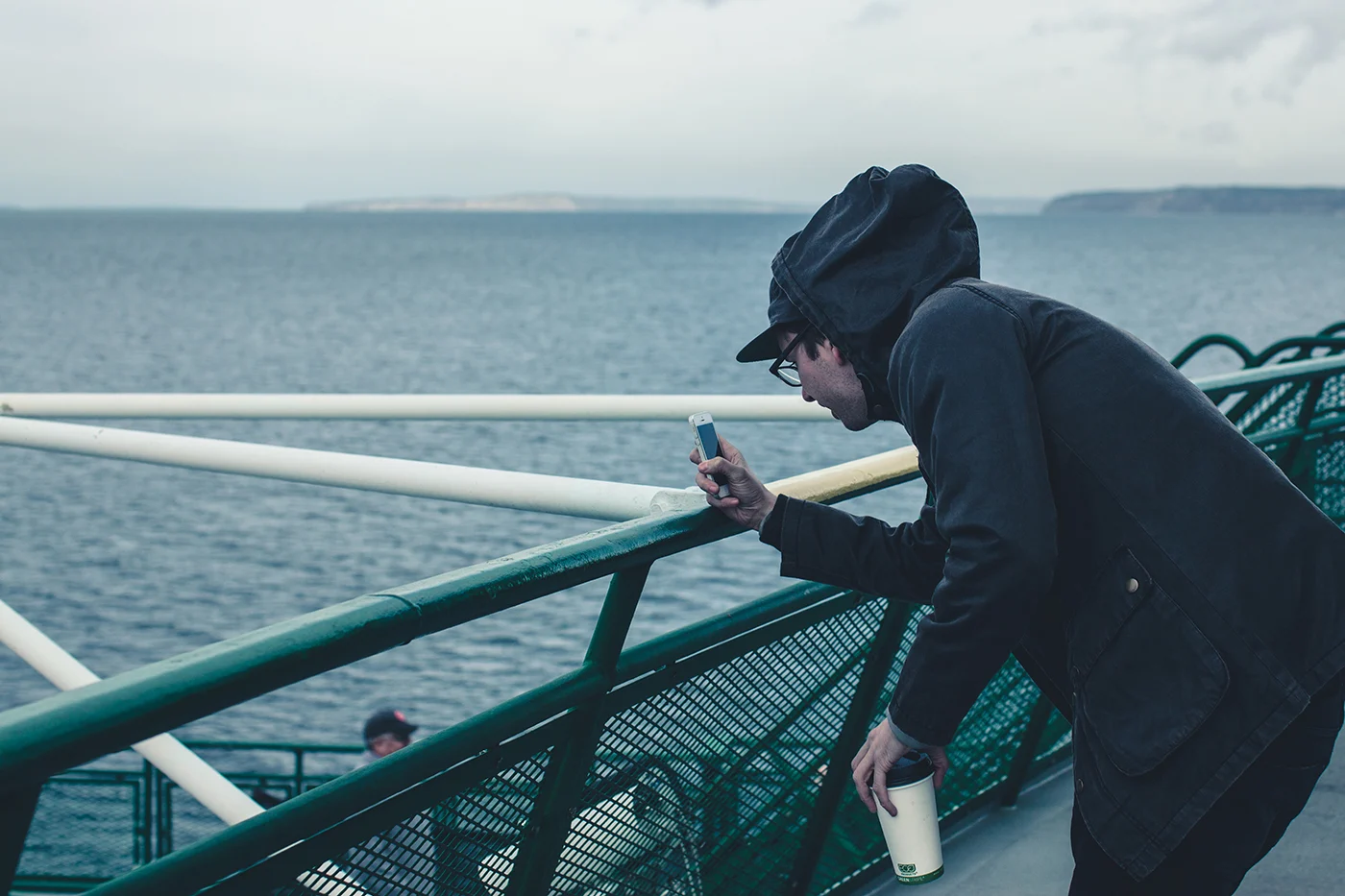 A man in a dark hood leaning on the rail of a ferry on a gray morning