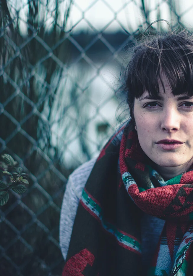 A young woman in a red plaid scarf by a chain-link fence on a rainy day
