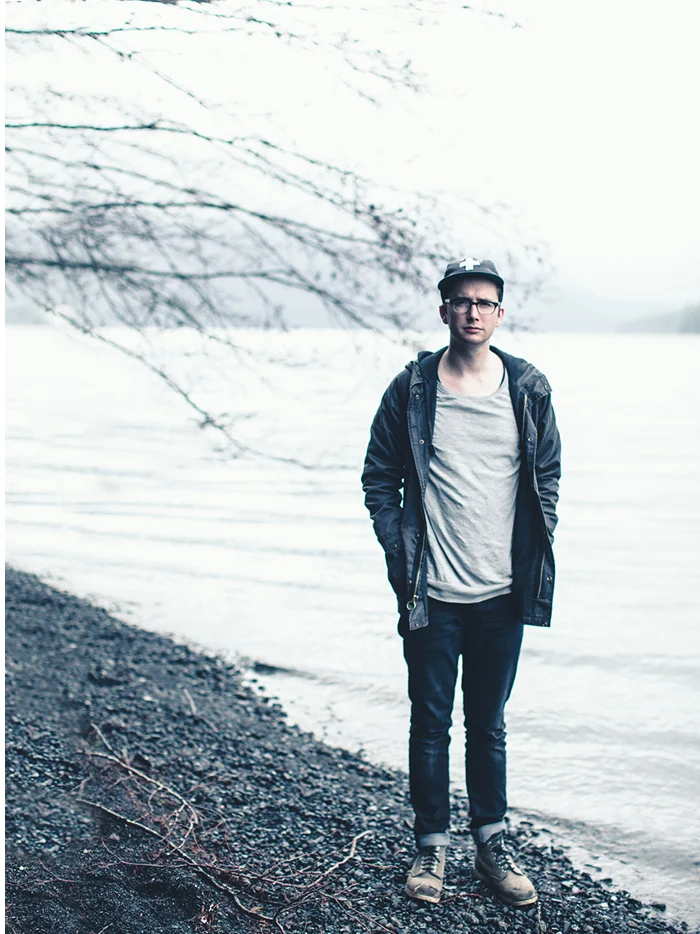 A young man standing on a pebble shore with the lake behind him
