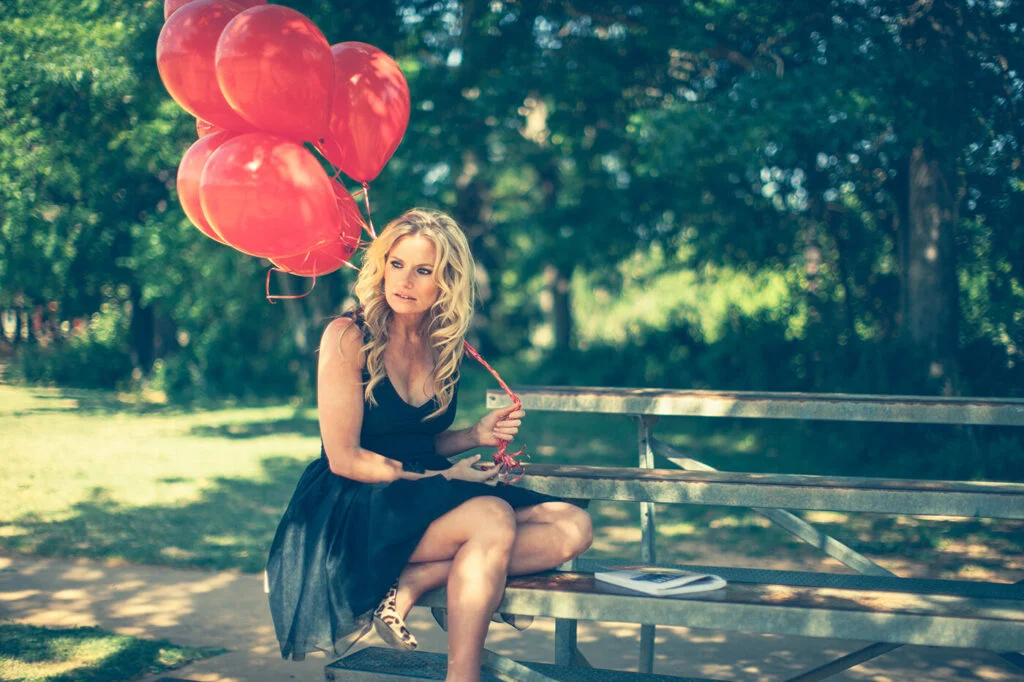 A woman in a black dress on a park bench holding a bunch of red balloons
