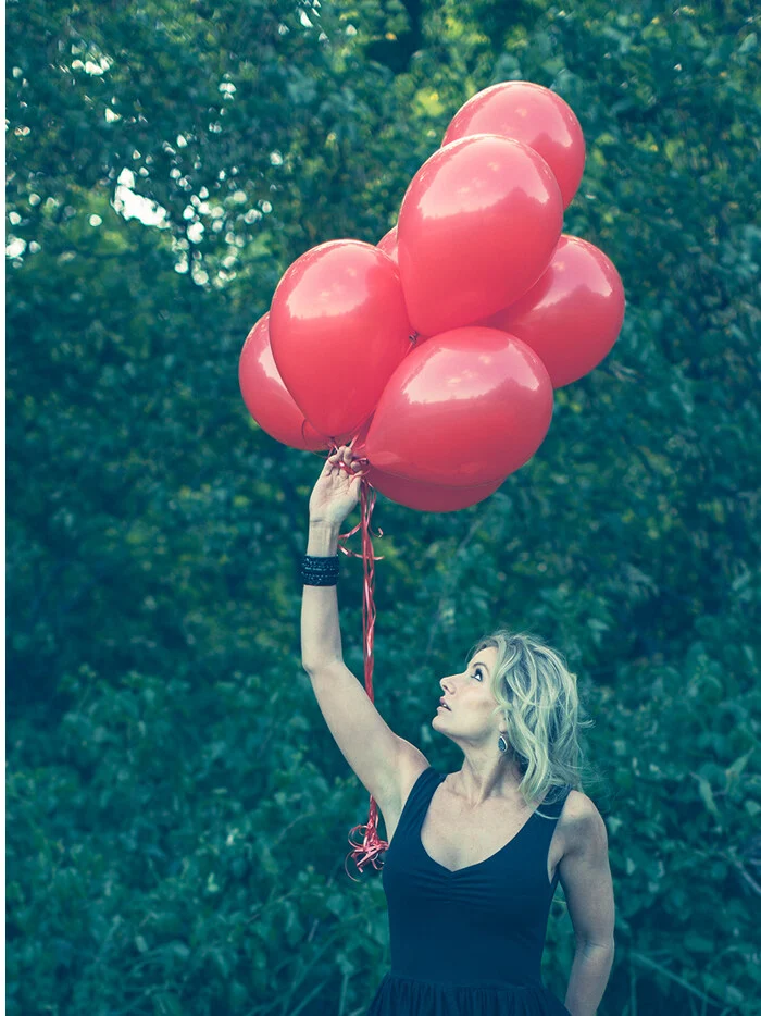 A woman holding a bunch of red balloons high above her head among green trees