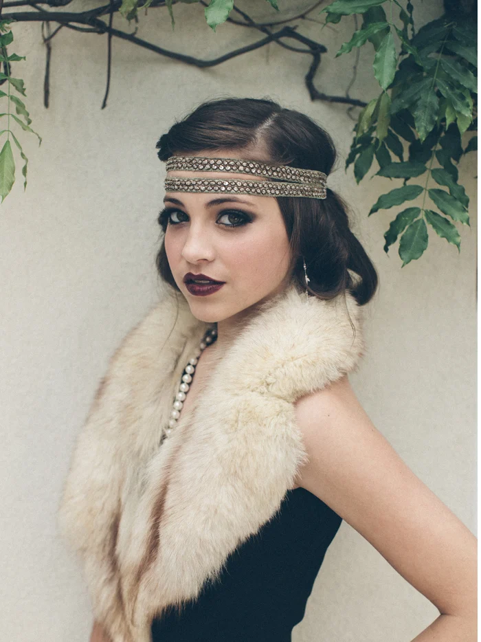 A woman in a beaded headband and a fur stole by an ivy-covered wall
